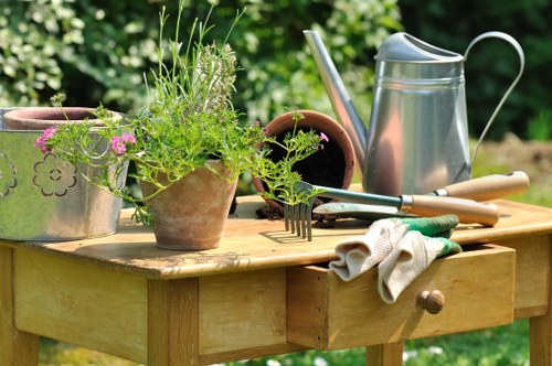 A wooden outdoor garden table in a lush green garden setting, displaying various gardening tools and potted plants. The table holds a silver watering can, a pair of gardening gloves, and multiple small terracotta pots, some filled with soil and others with young plant growth or flowering plants, including purple and pink blossoms. The surface of the table is slightly weathered, indicating regular outdoor use, with a natural wood grain visible. In the background, dense foliage and garden greenery provide a peaceful outdoor environment, suggesting a well-maintained garden space suitable for planting and outdoor maintenance activities, characteristic of gardening services in Norbiton or nearby areas.