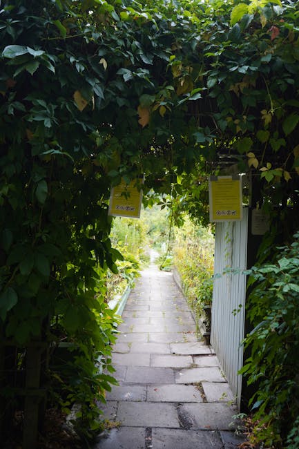 A narrow garden pathway paved with rectangular grey stones, leading through a lush, overgrown green archway formed by dense leafy foliage and branches. On the right side, a partially visible white woo