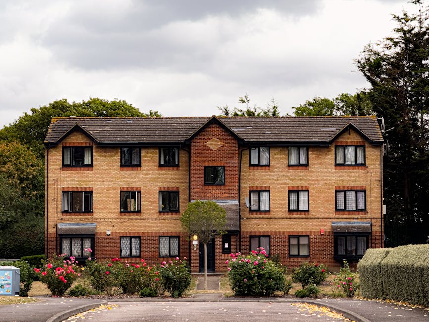 The image shows the exterior of a red brick residential building with a tiled roof and white-framed windows, some with closed white blinds. A lush, well-maintained shrub with dense green foliage and c